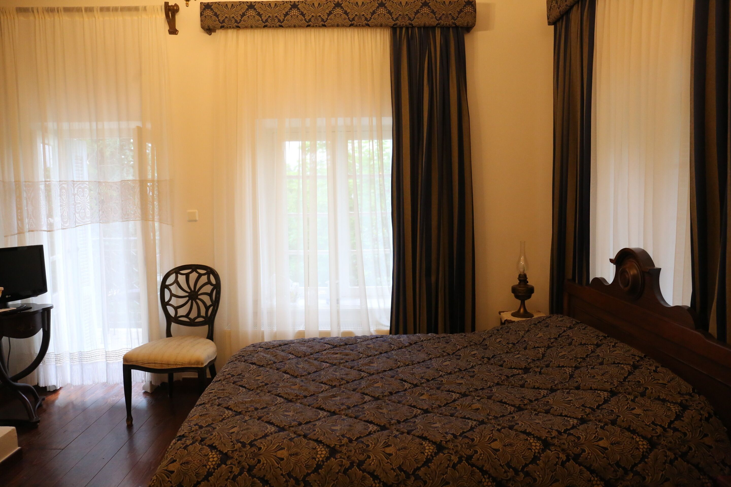 A bedroom with a large bed, a chair, a desk with a computer, and a window with curtains.