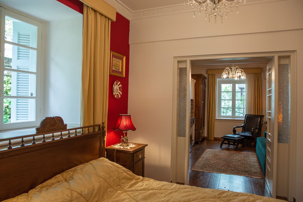 Habitación de hotel con cama, mesita de noche con lámpara, pared roja, ventana con contraventanas y vista al pasillo.