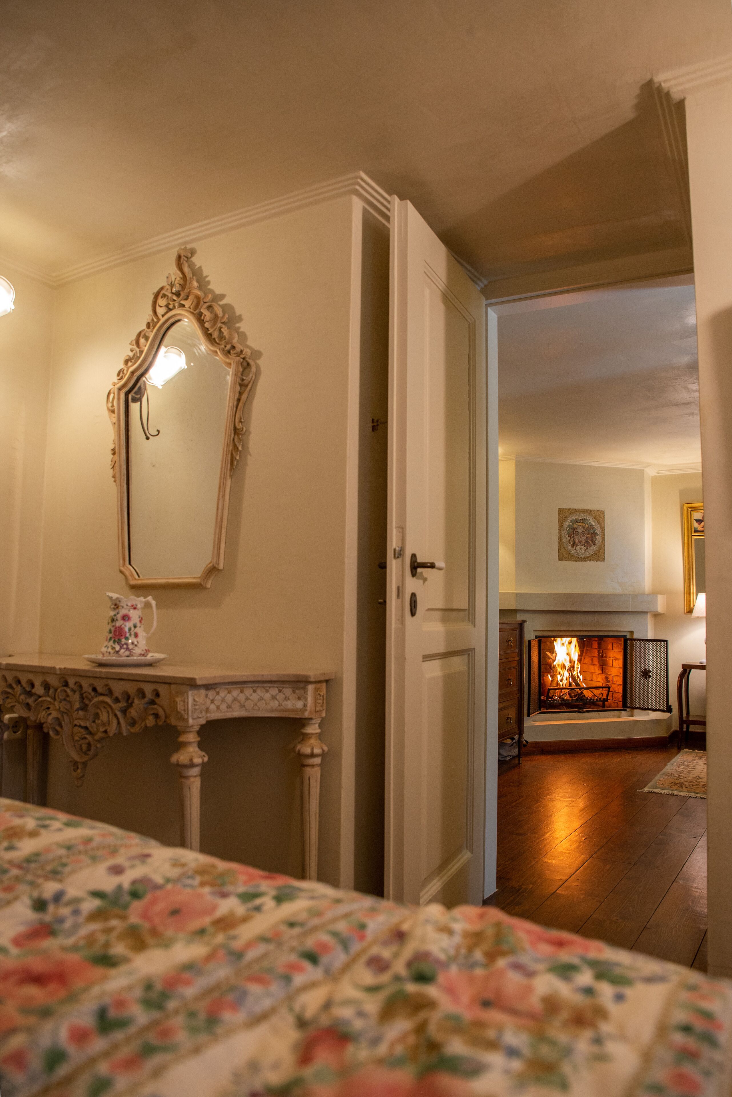 A room with a bed, a fireplace, a mirror, and a door leading to another room.