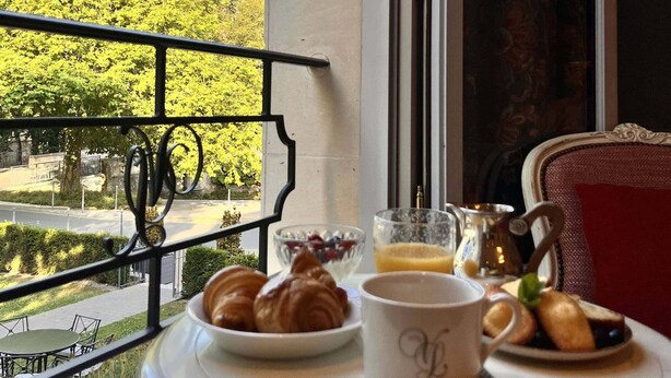A table with croissants, a cup, and a glass of orange juice on a balcony with a view of a church and trees. - Villa Lara Hôtel (Bayeux)