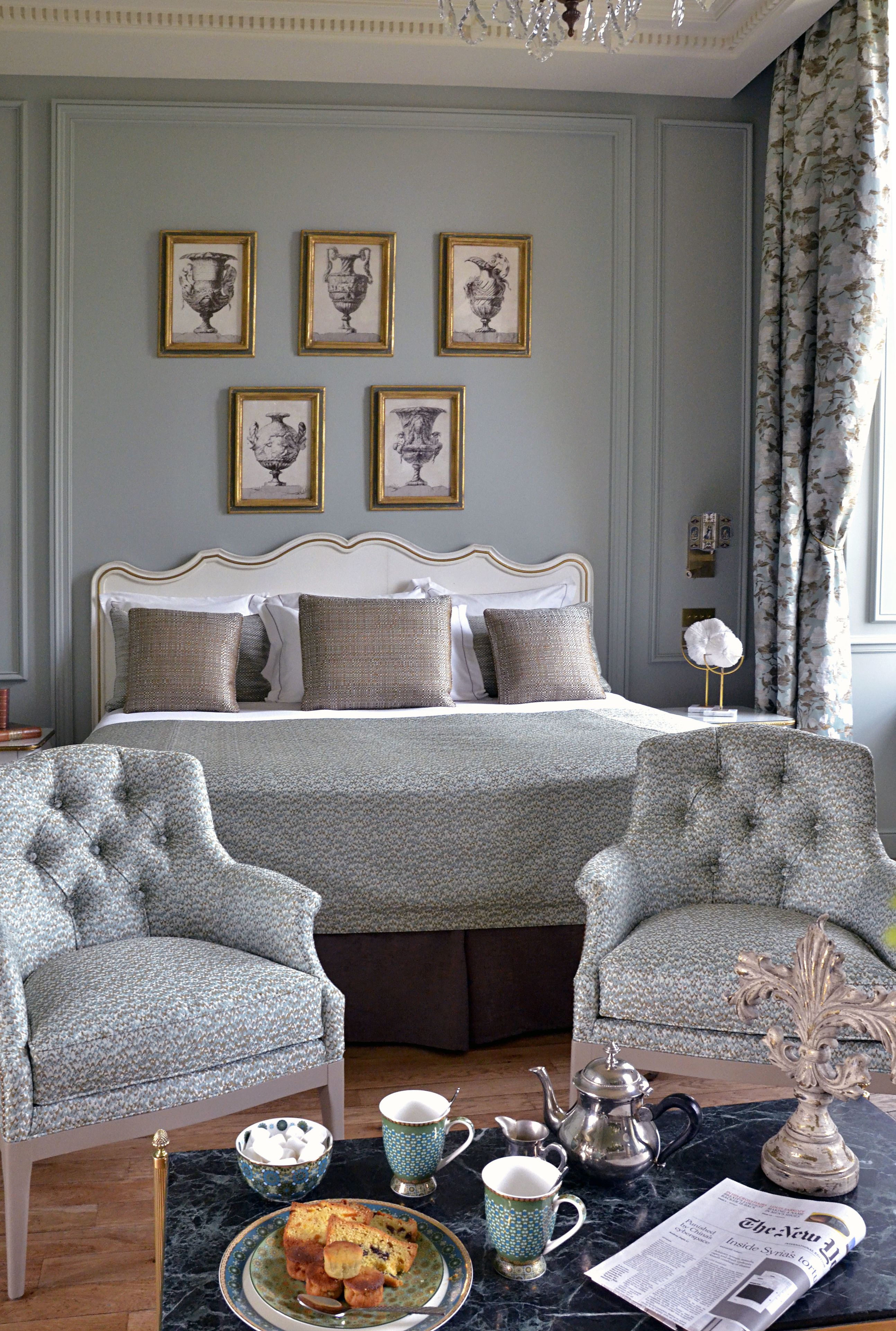 A neatly arranged bedroom with a bed, two armchairs, a coffee table with breakfast items, and framed artwork on the wall.
