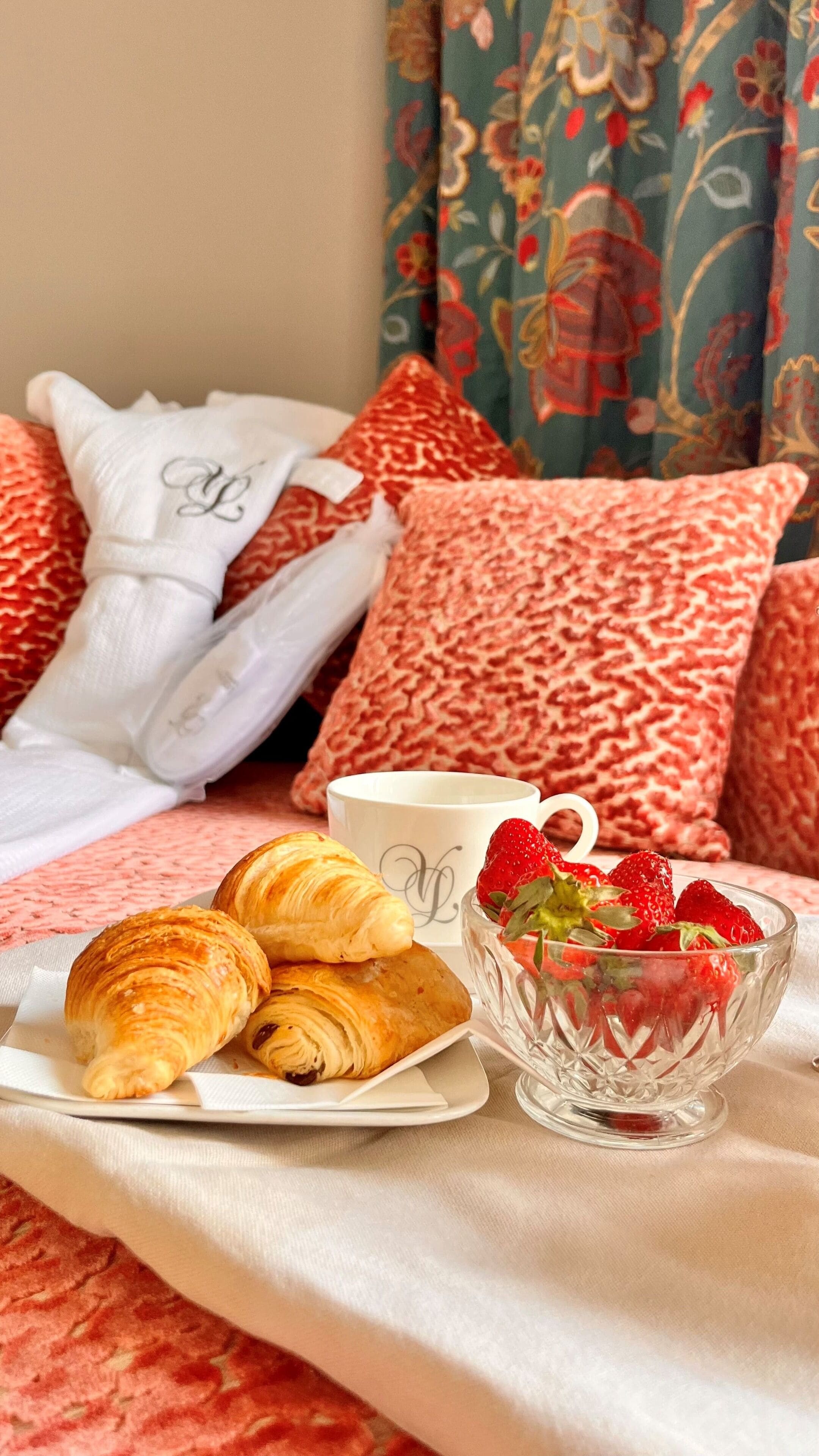 A breakfast setup with croissants, strawberries, and a cup on a bed with patterned pillows and a floral curtain.