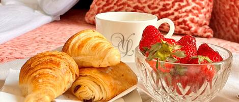 A breakfast setup with croissants, strawberries, and a cup on a bed with patterned pillows and a floral curtain.