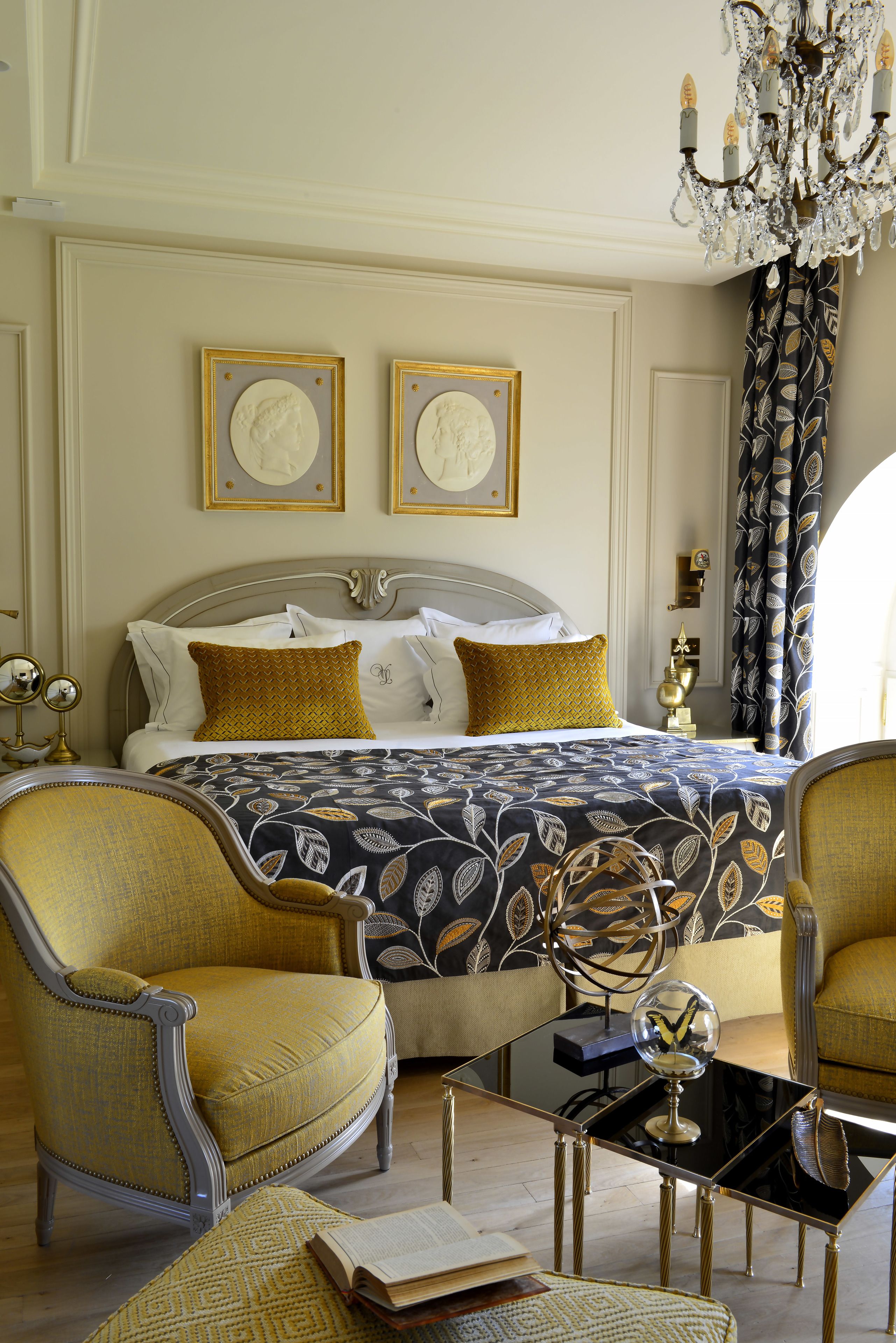 Un dormitorio elegante con una cama grande, dos retratos enmarcados, una araña y muebles clásicos.