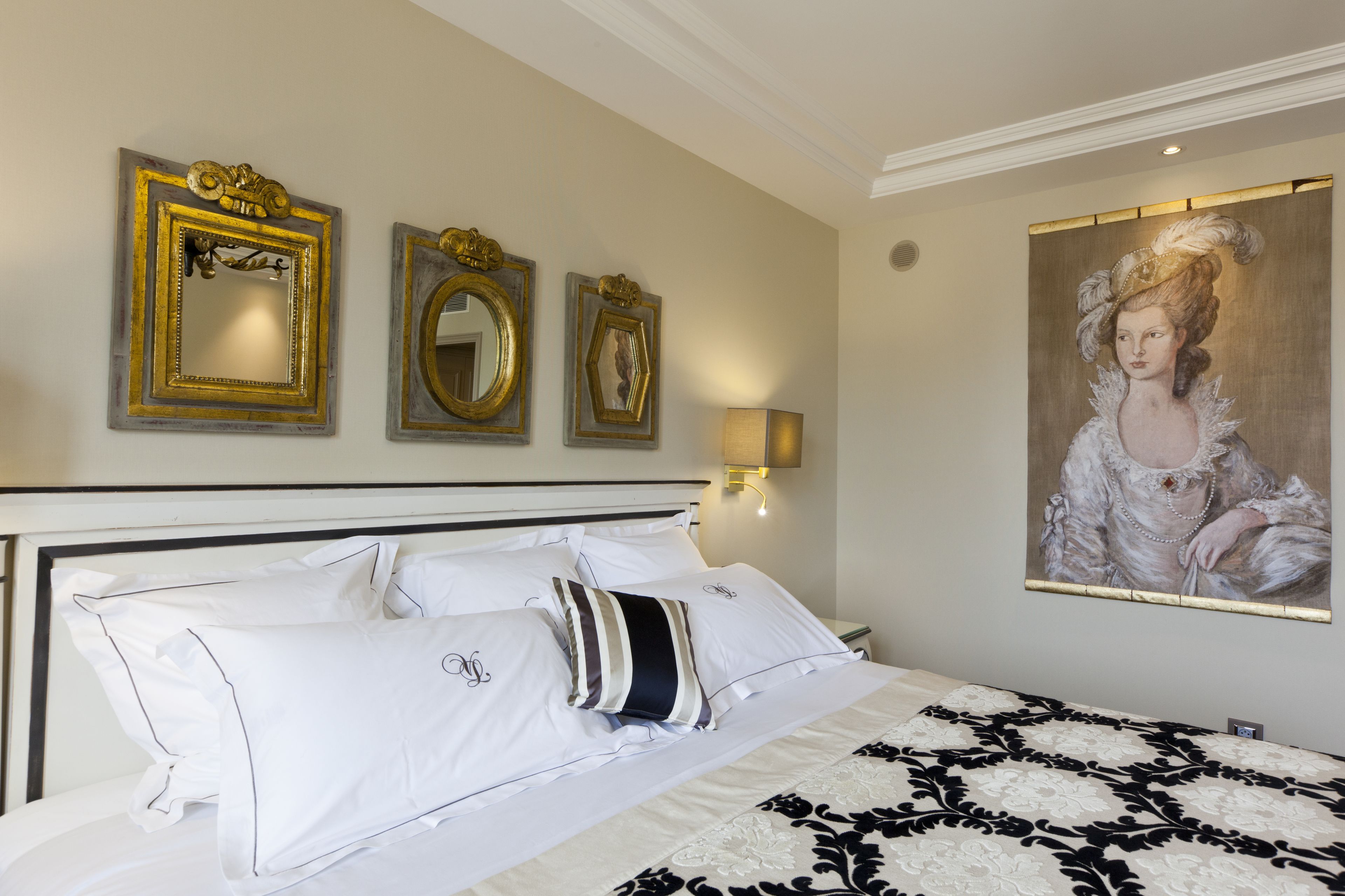 Un lit bien fait, avec du linge de lit blanc et une couvre-lit à motifs noir et blanc. Trois miroirs encadrés sont accrochés au mur au-dessus du lit, et un tableau représentant une femme en blanc est suspendu sur le côté droit.
