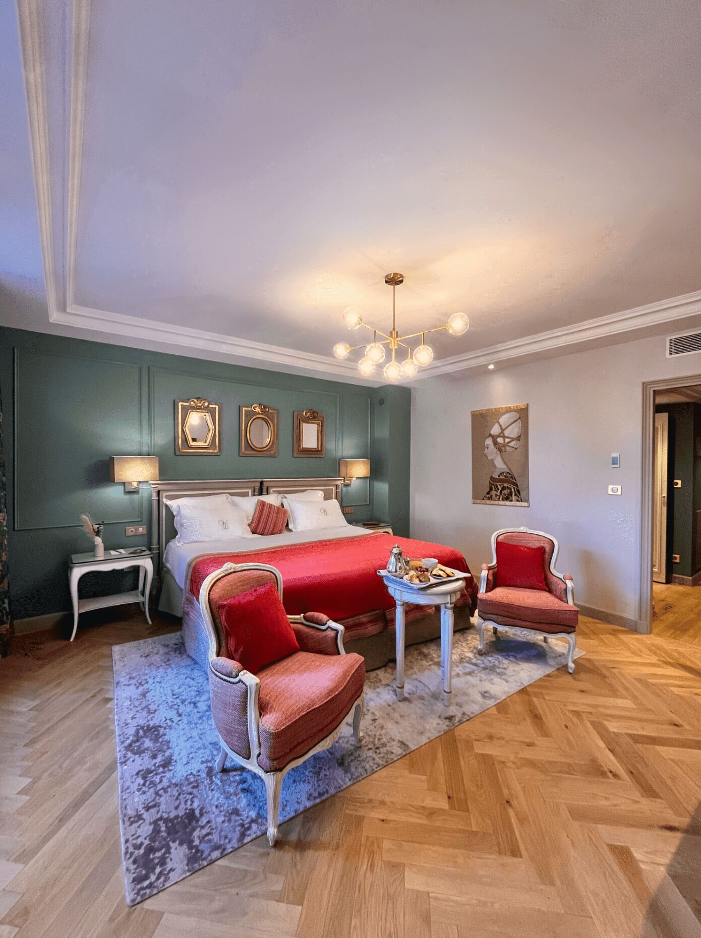 A hotel room with a large bed, two armchairs, a small table with breakfast, and a chandelier.