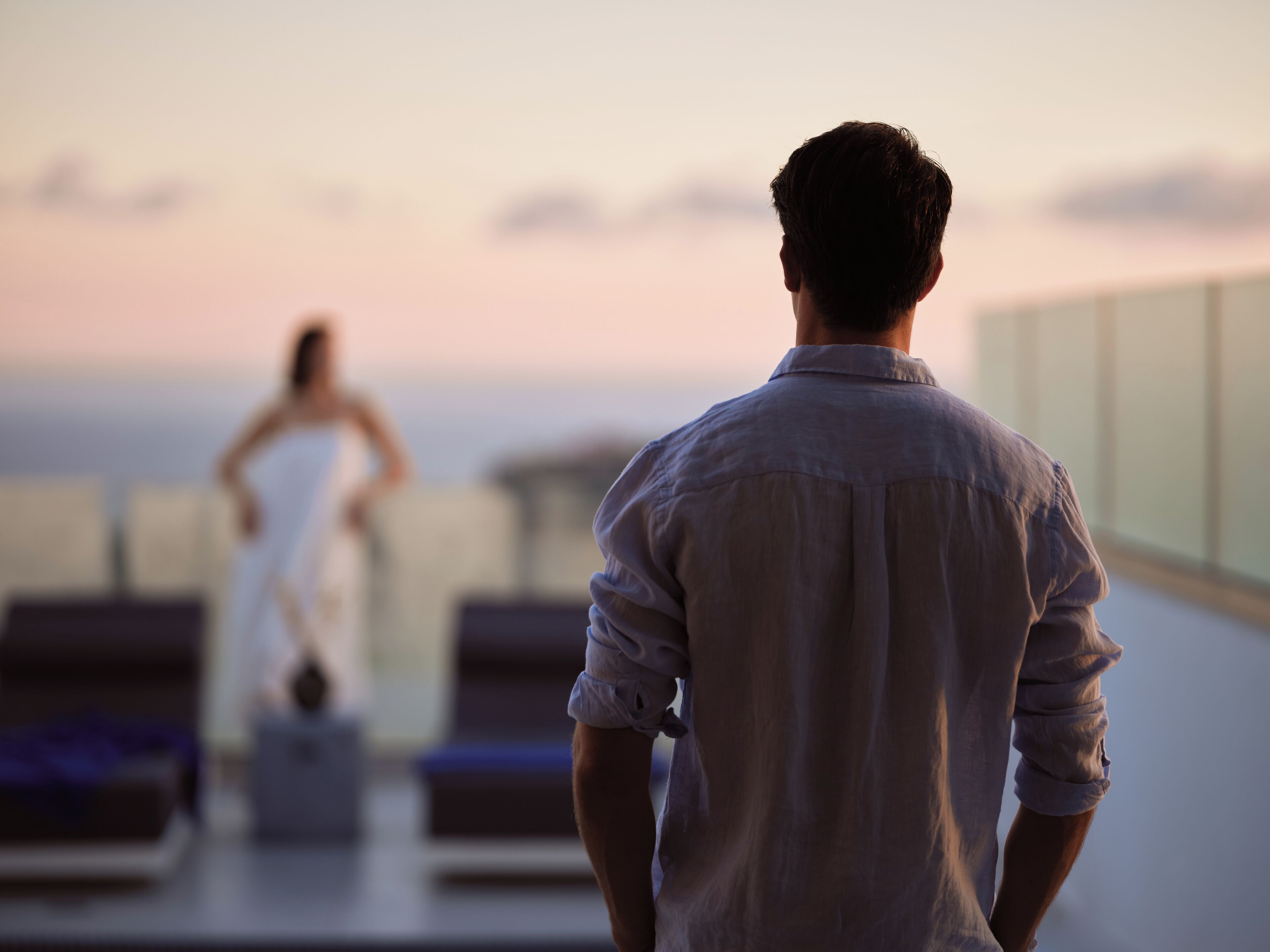 Un hombre con camisa blanca está de pie en un balcón, observando a una mujer con vestido blanco.