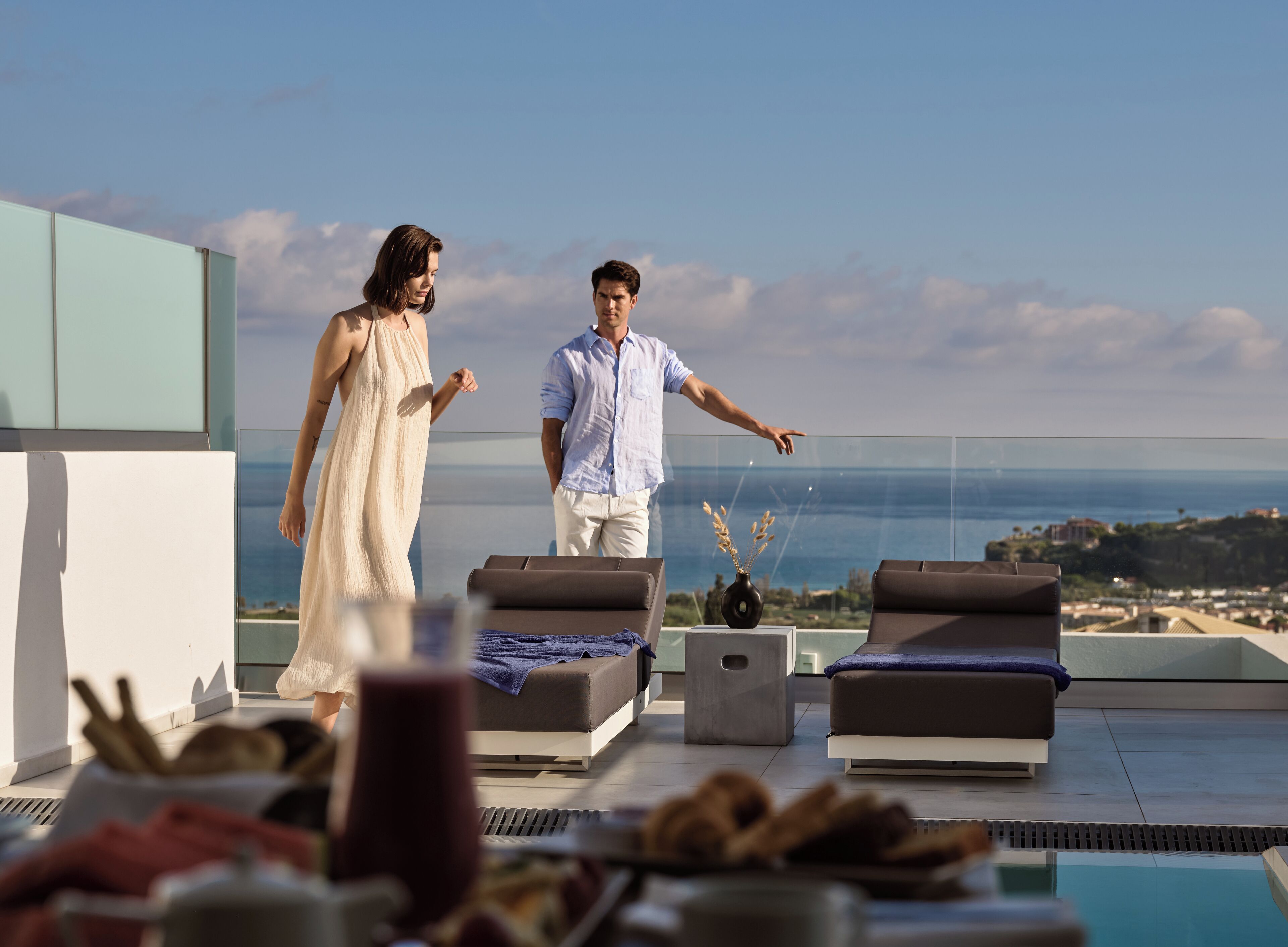 Um casal caminhando em um terraço com piscina e espreguiçadeiras, com vista para a paisagem costeira.