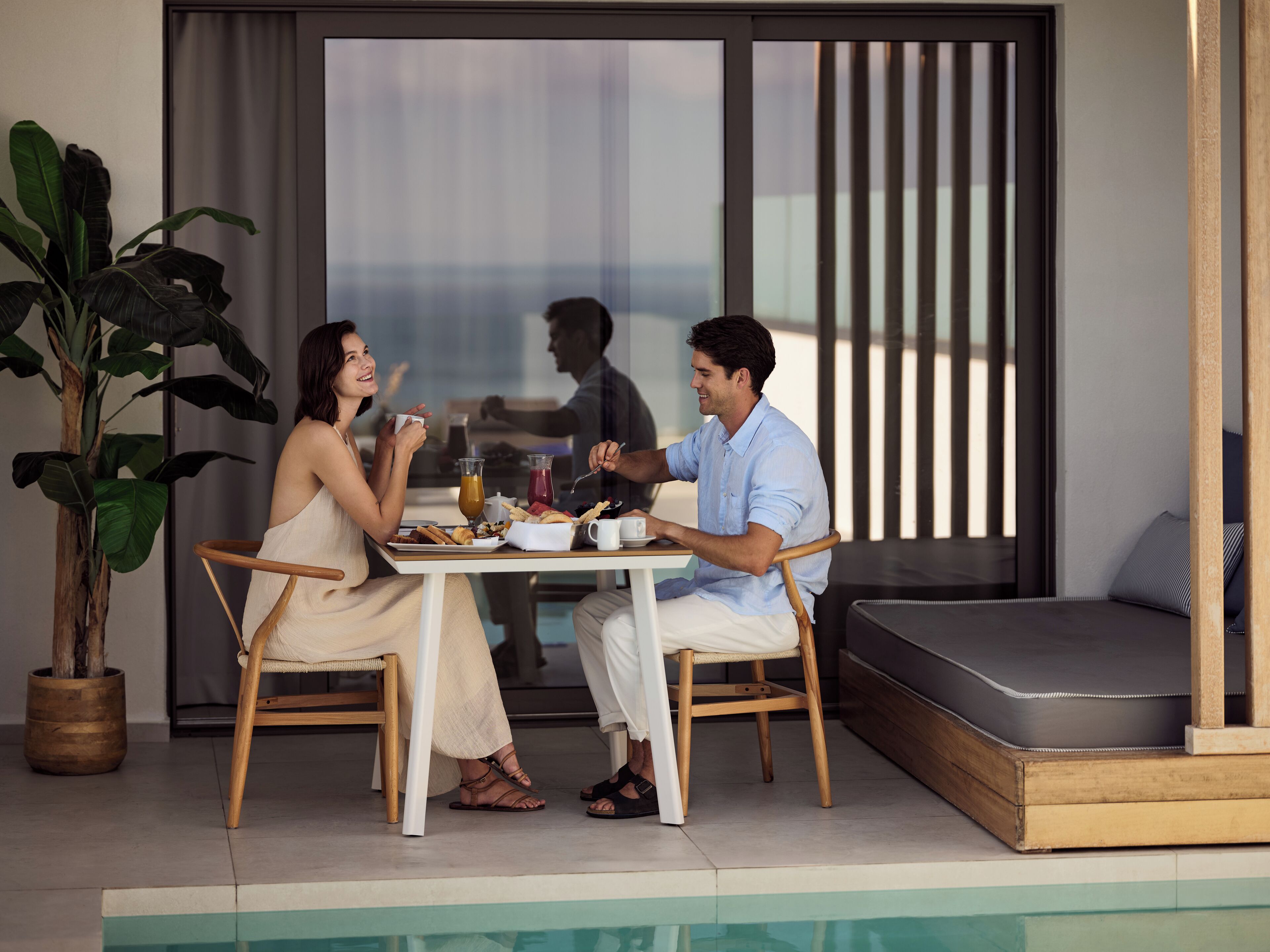 A dining area with a table set for two, a potted plant, and a view of the outdoors.