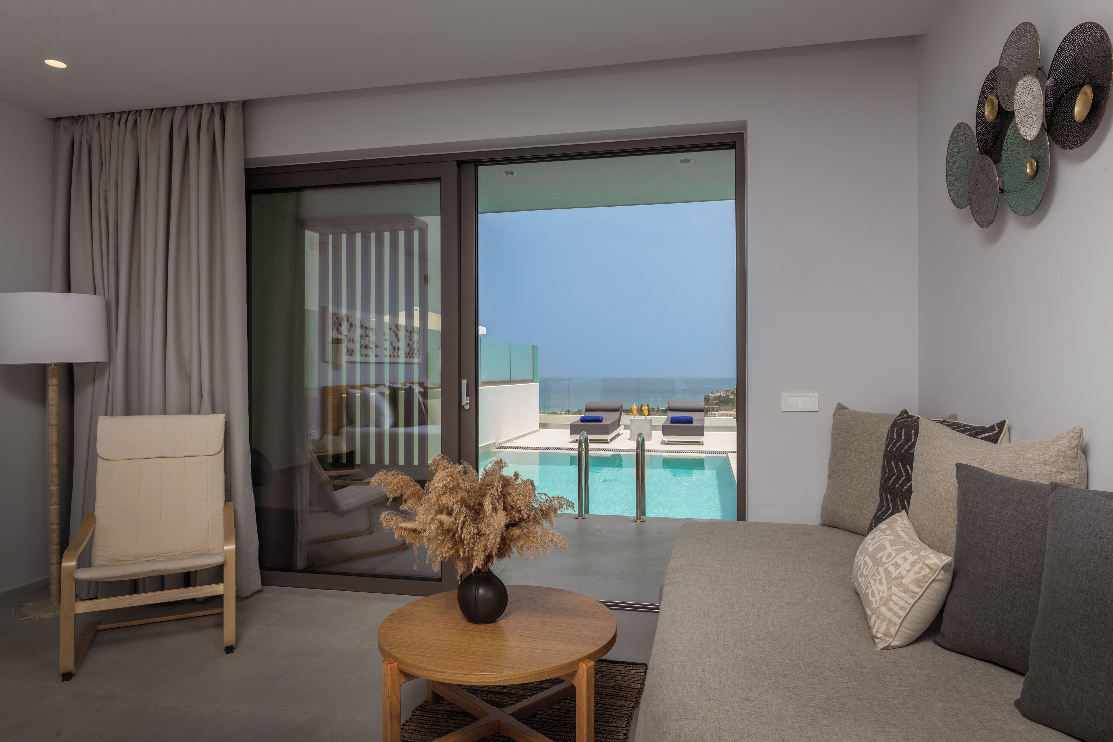 Un salon moderne comprenant un canapé, un fauteuil, une petite table avec un vase, et une vue sur une piscine et un espace de détente, le tout visible à travers une porte vitrée.