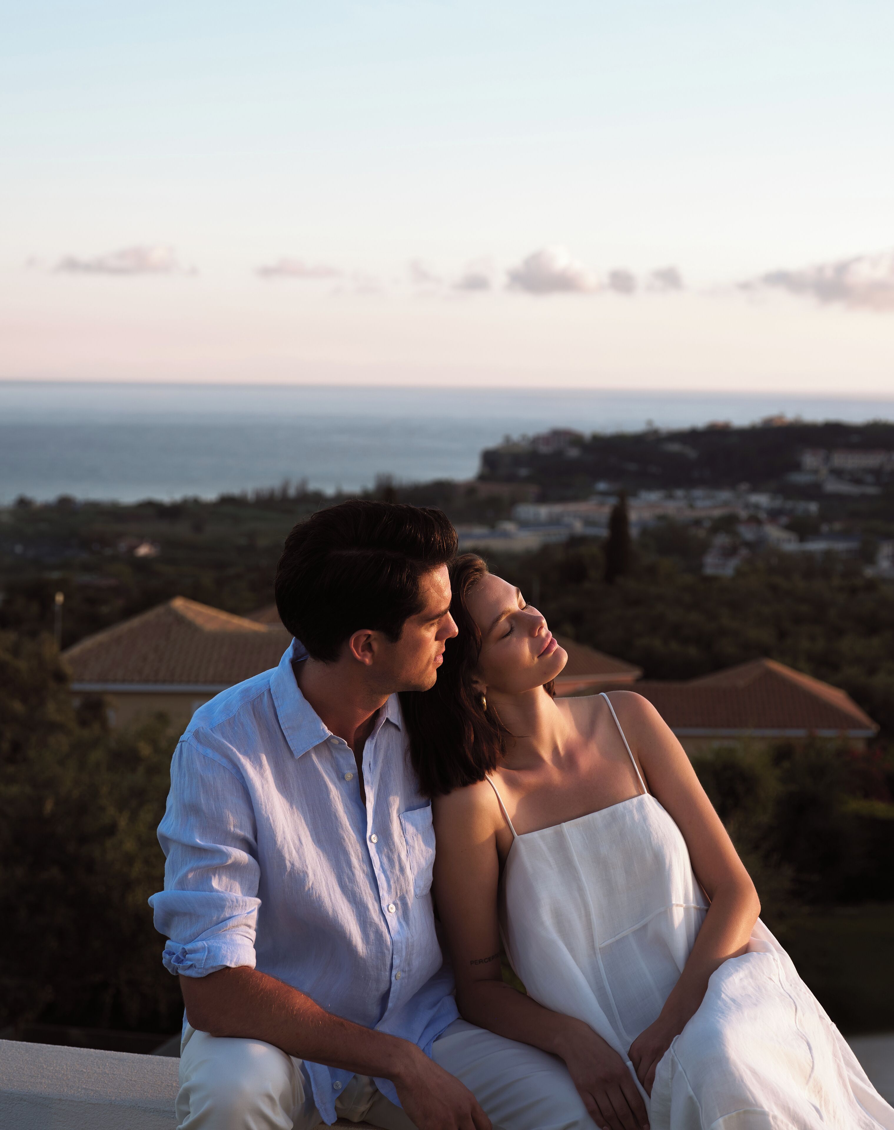Una pareja sentada en un balcón con vistas a la costa y casas al fondo.