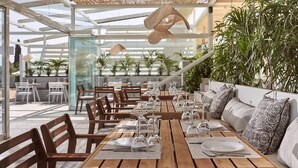 A modern outdoor dining area with wooden tables and chairs, glass partitions, and woven ceiling lamps.