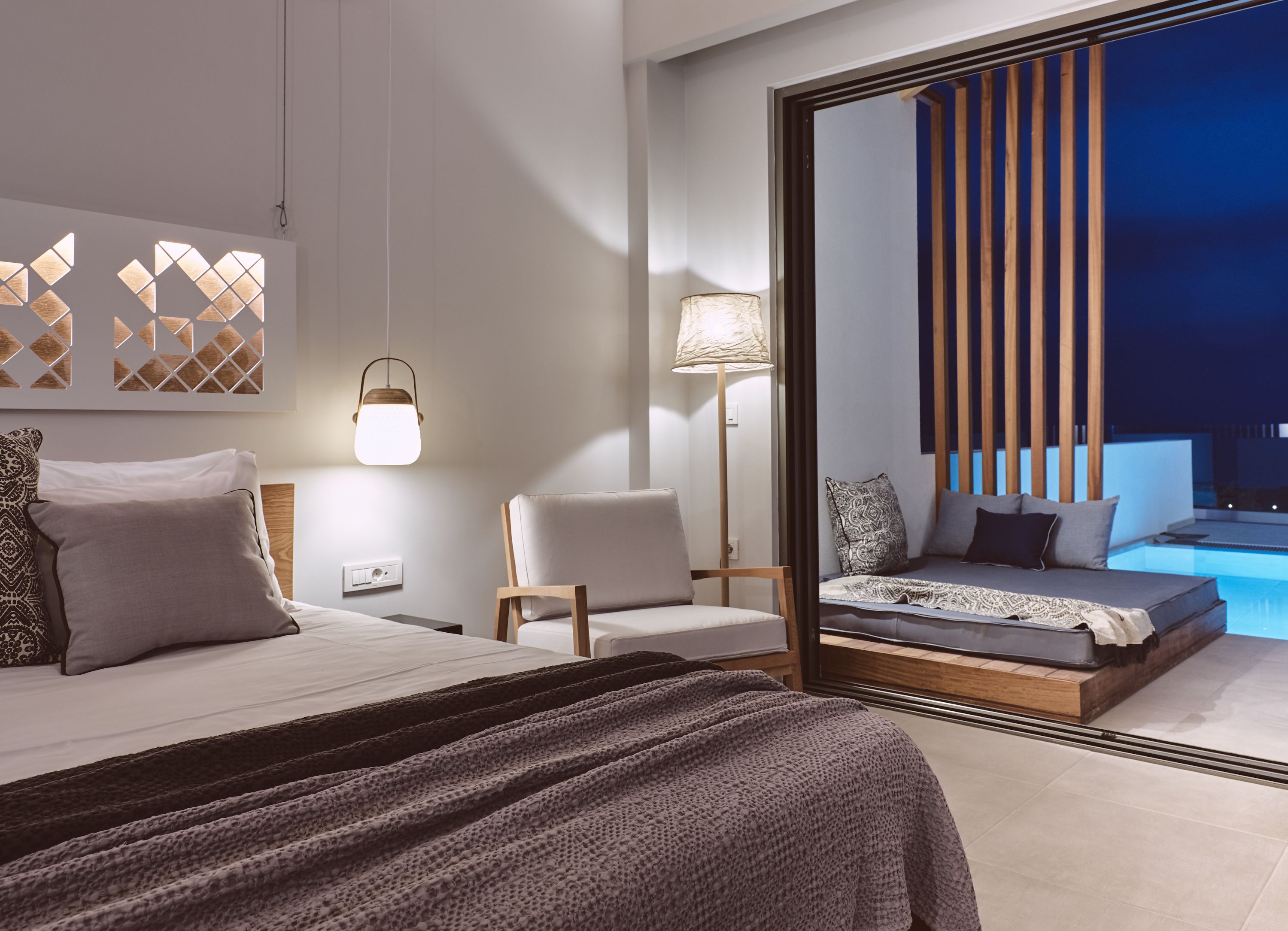 A modern hotel room with a bed, a chair, a bedside table, a lamp, and a view of a pool at night.