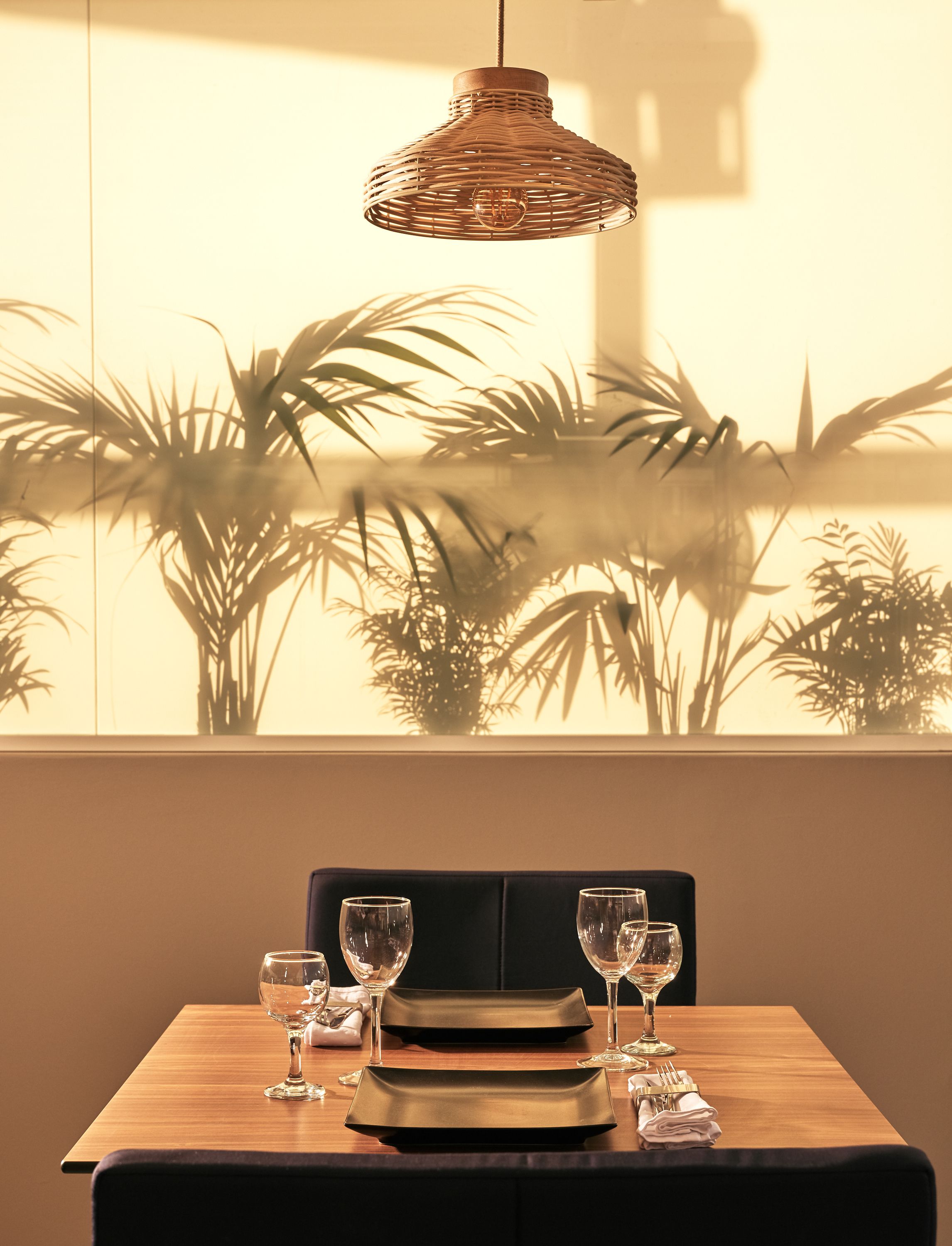 Een eettafel gedekt met borden, glazen en bestek, met een rieten hanglamp erboven en uitzicht op palmbomen door een raam.