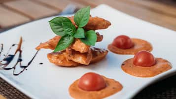 Ein weißer, quadratischer Teller mit Speisen, darunter ein Stapel knuspriger Snacks, ein Basilikumblatt und zwei kleine Tomatenstückchen auf einer strukturierten Oberfläche.