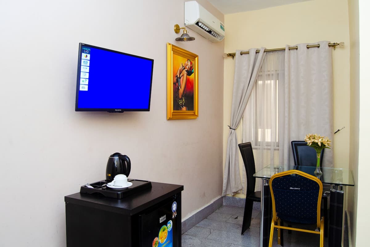 A hotel room with a flat-screen TV, a framed picture, a coffee maker, a glass table, a chair, and a vase with flowers.