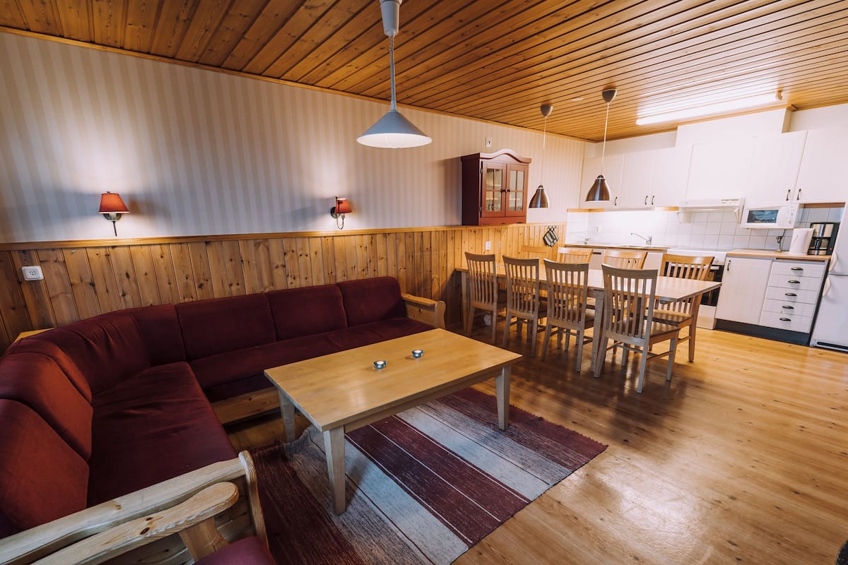 A cozy room with a wooden ceiling, a red sofa, a wooden dining table, and a kitchen area with white cabinets and a microwave.