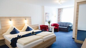 A modern hotel room with a wooden bed, a red armchair, a grey sofa, and a small table with a vase of flowers. - Hotel Pellmühle (Jever)