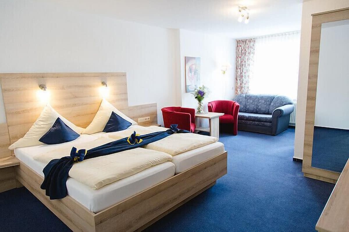 A modern hotel room with a wooden bed, a red armchair, a grey sofa, and a small table with a vase of flowers.
