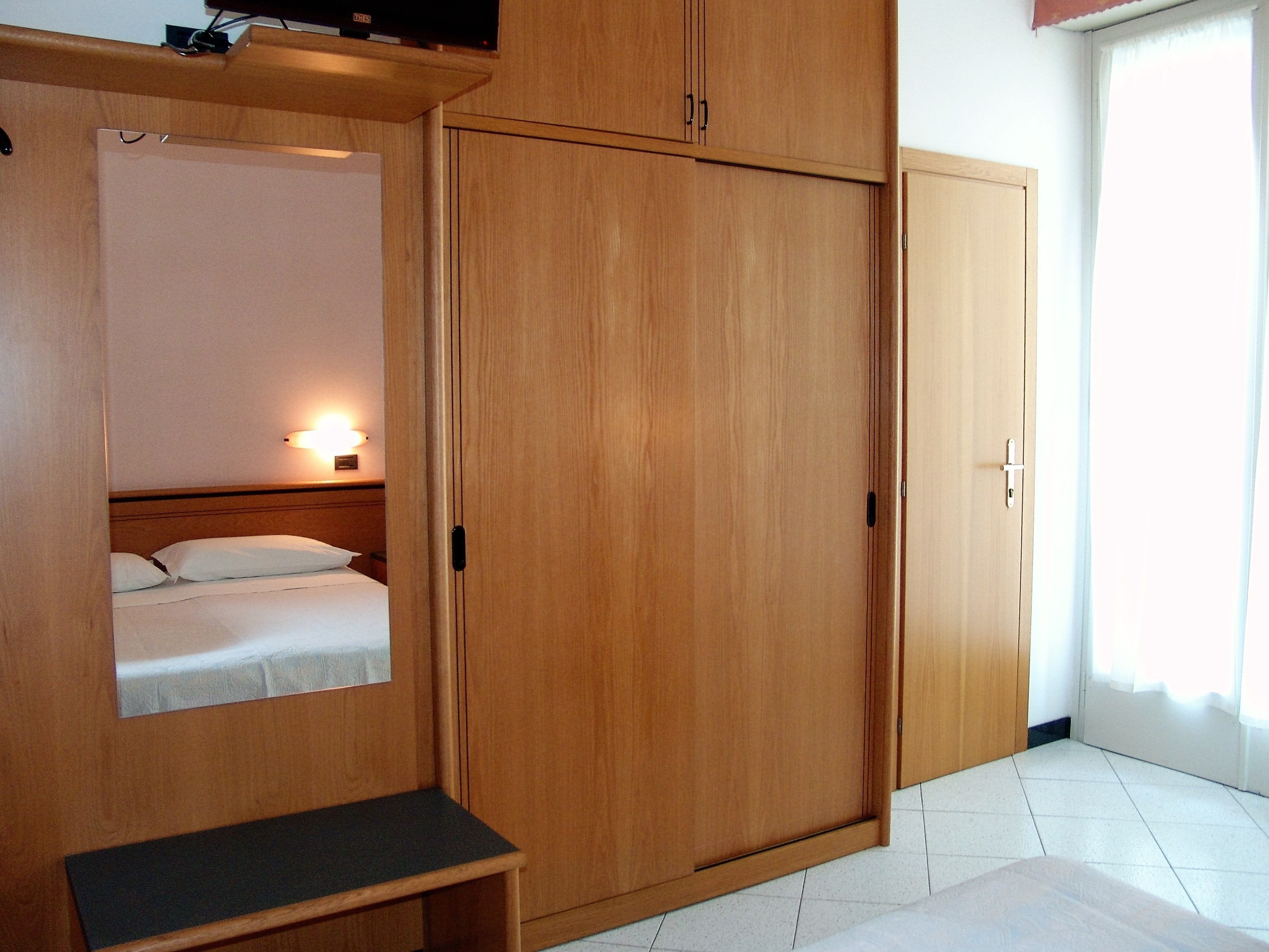A room with a wooden wardrobe, a bed with white bedding, a bedside table, and a wall-mounted light.