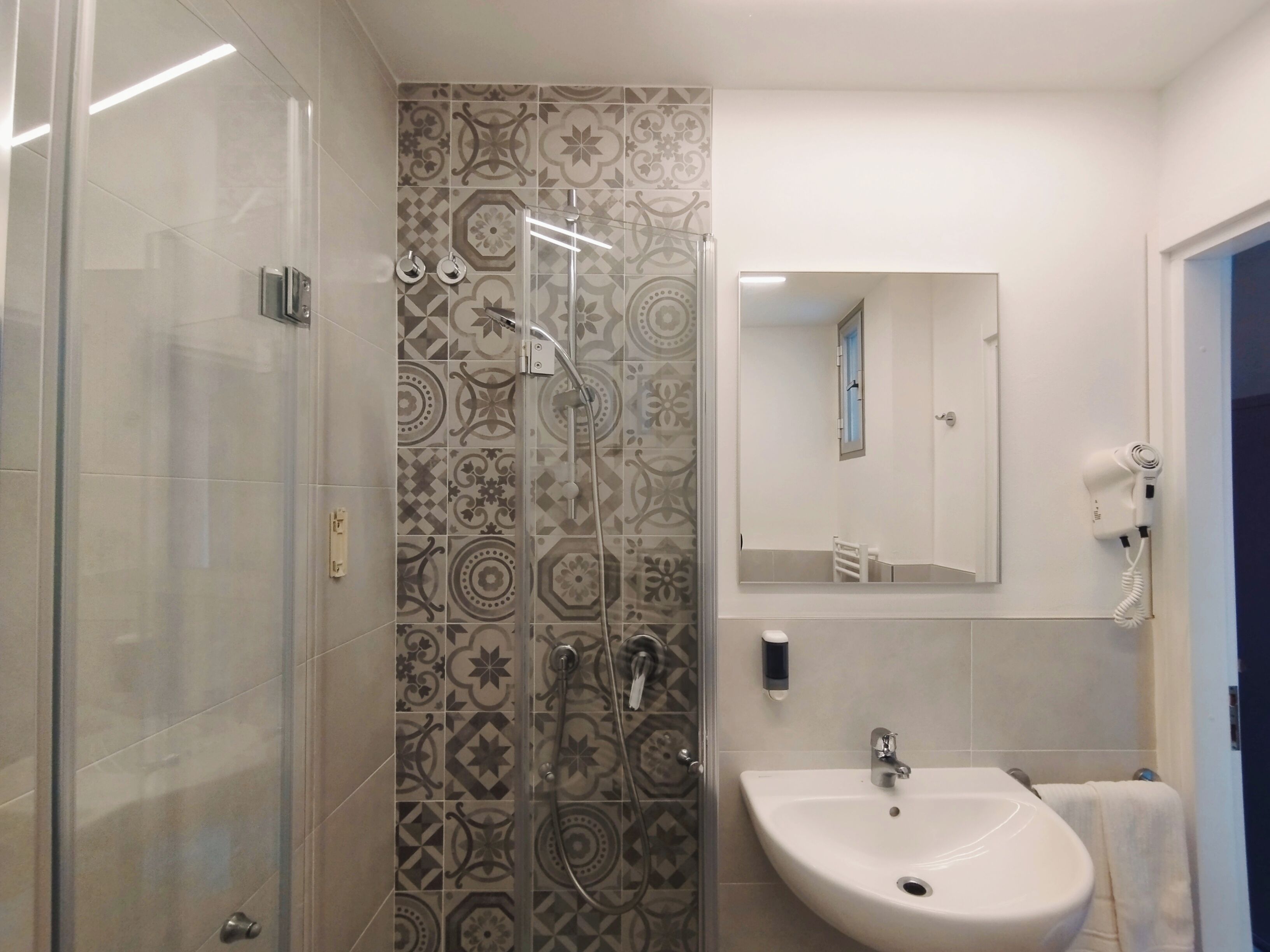 A modern bathroom with a glass shower enclosure, patterned tiles, a white sink, and a mirror with a built-in light.