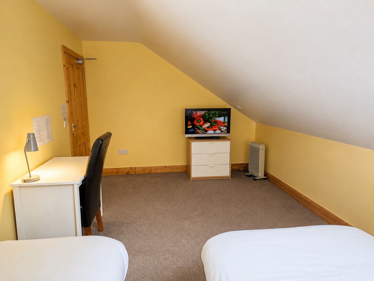 Une petite chambre d’hôtel composée d’une seule pièce, avec un plafond en pente, un bureau avec une lampe, une chaise, un téléviseur posé sur une commode et deux lits.