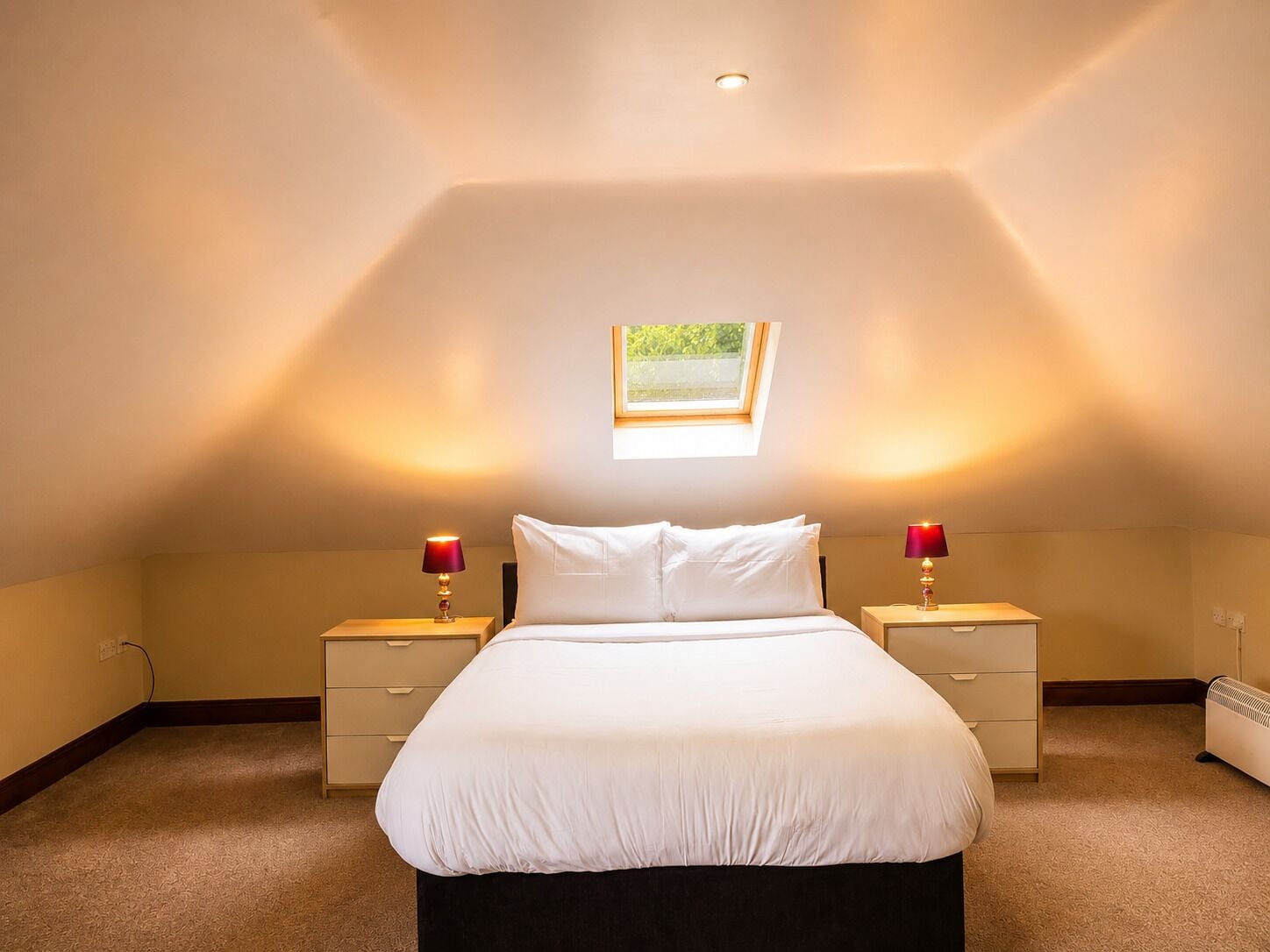 A bedroom with a skylight, a bed with white linens, two bedside tables with lamps, and a radiator.