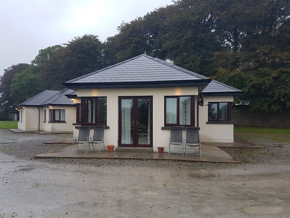 A small, single-story building with a tiled roof, a front door, and two windows with curtains. There are two chairs outside the building. - Moorepark West House (Fermoy)