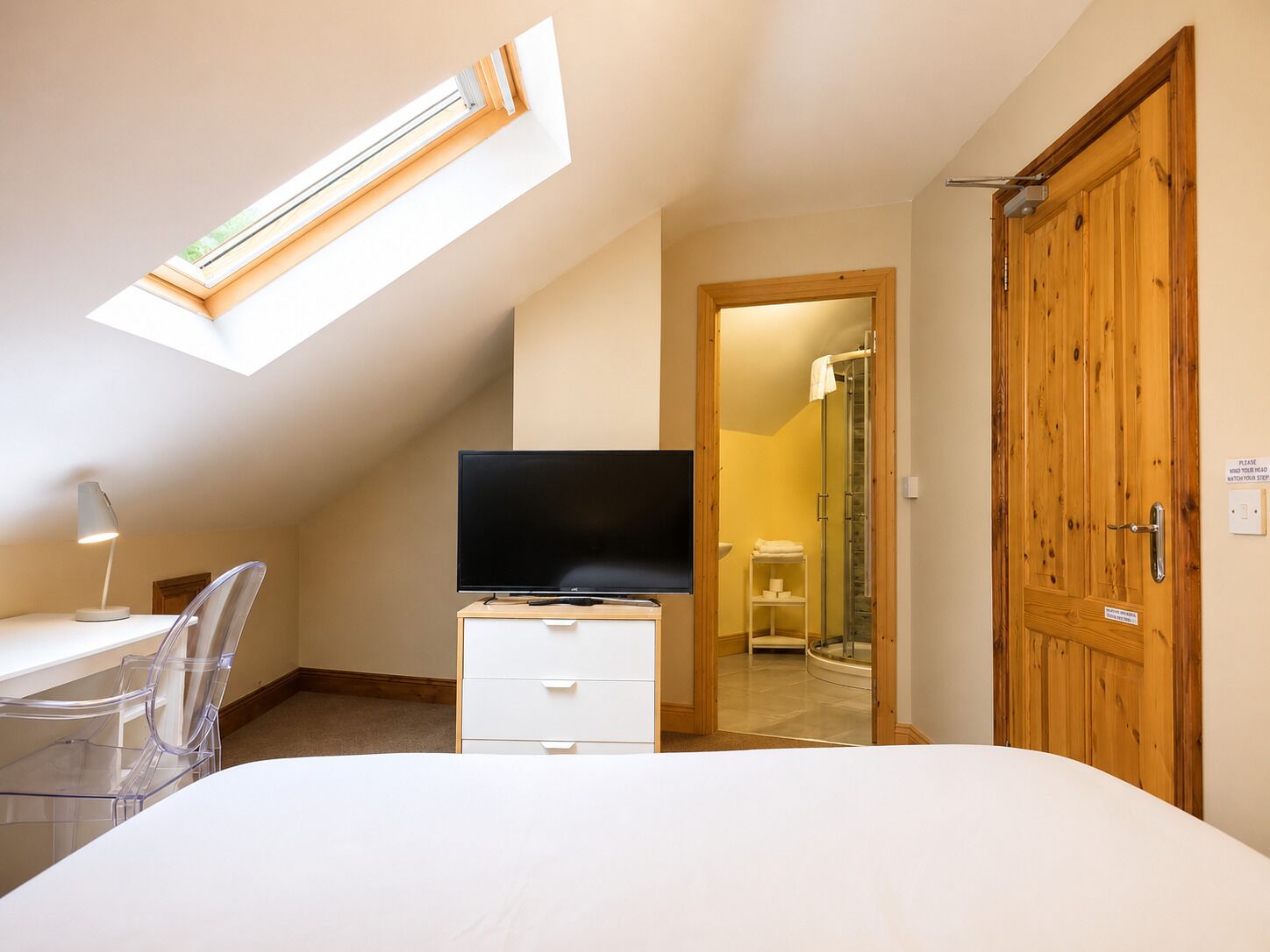 Ein Schlafzimmer mit Oberlicht, einem Fernseher auf einer Kommode, einem Schreibtisch mit Stuhl und einer Holztür.