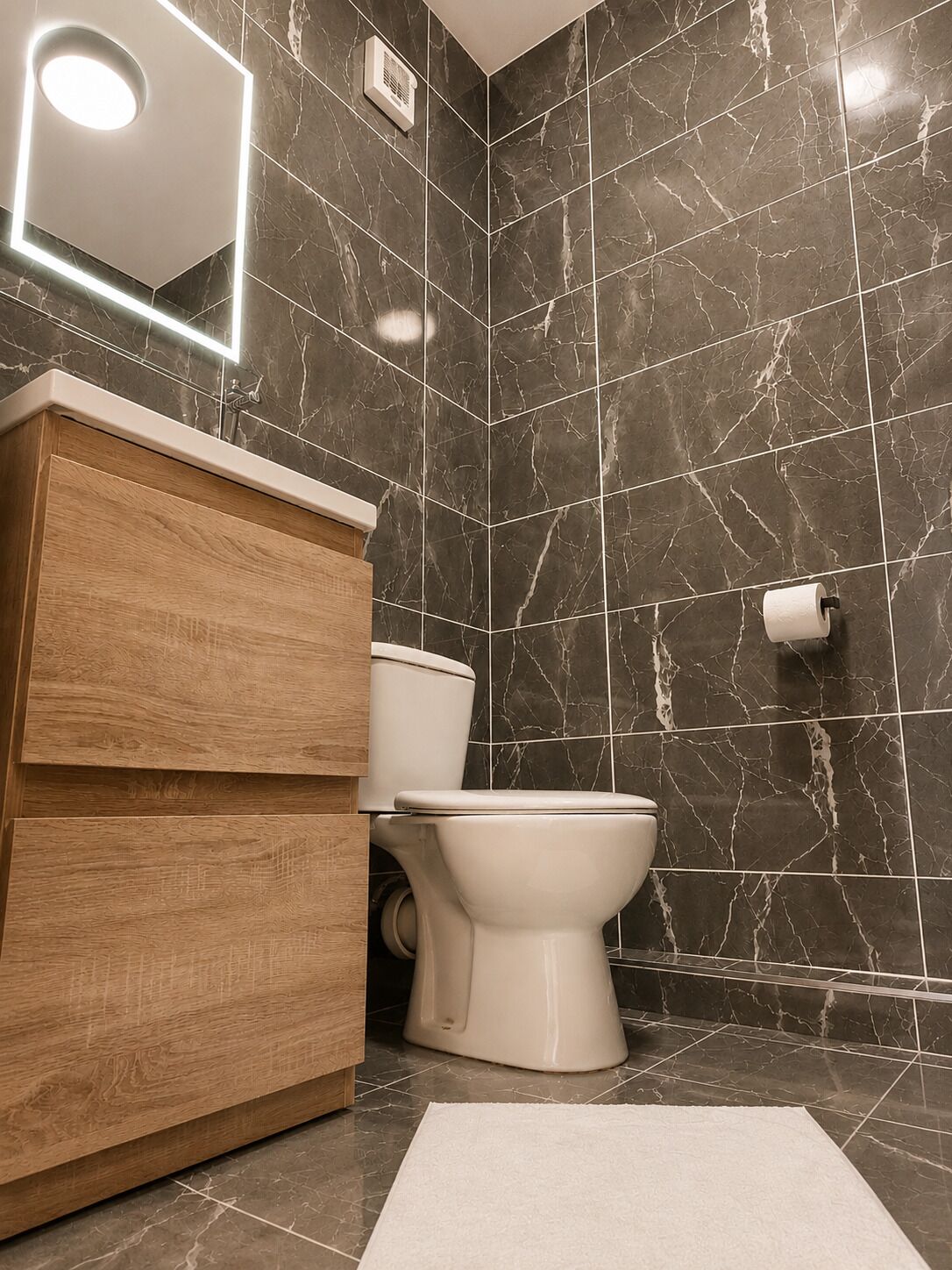 A modern bathroom with a marble wall, a wooden vanity, a toilet, and a roll of toilet paper on the wall.
