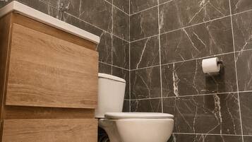 A modern bathroom with a marble wall, a wooden vanity, a toilet, and a roll of toilet paper on the wall.