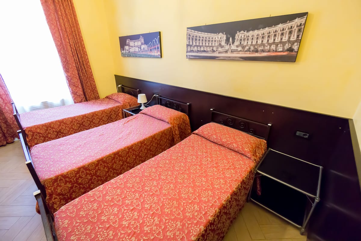A hotel room with three single beds, each with a patterned bedspread. There are two framed pictures on the wall, one depicting a bridge and the other a building.