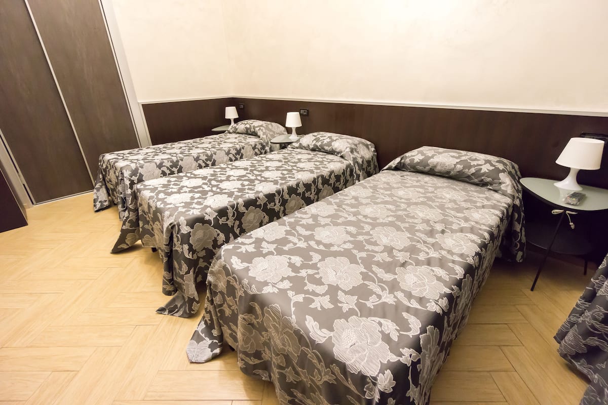 A hotel room with three beds, each with a floral-patterned bedspread, a small table with a lamp, and a wooden floor.