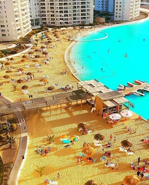 Un complejo turístico en la playa con varios edificios, una playa de arena y una piscina azul cristalina.