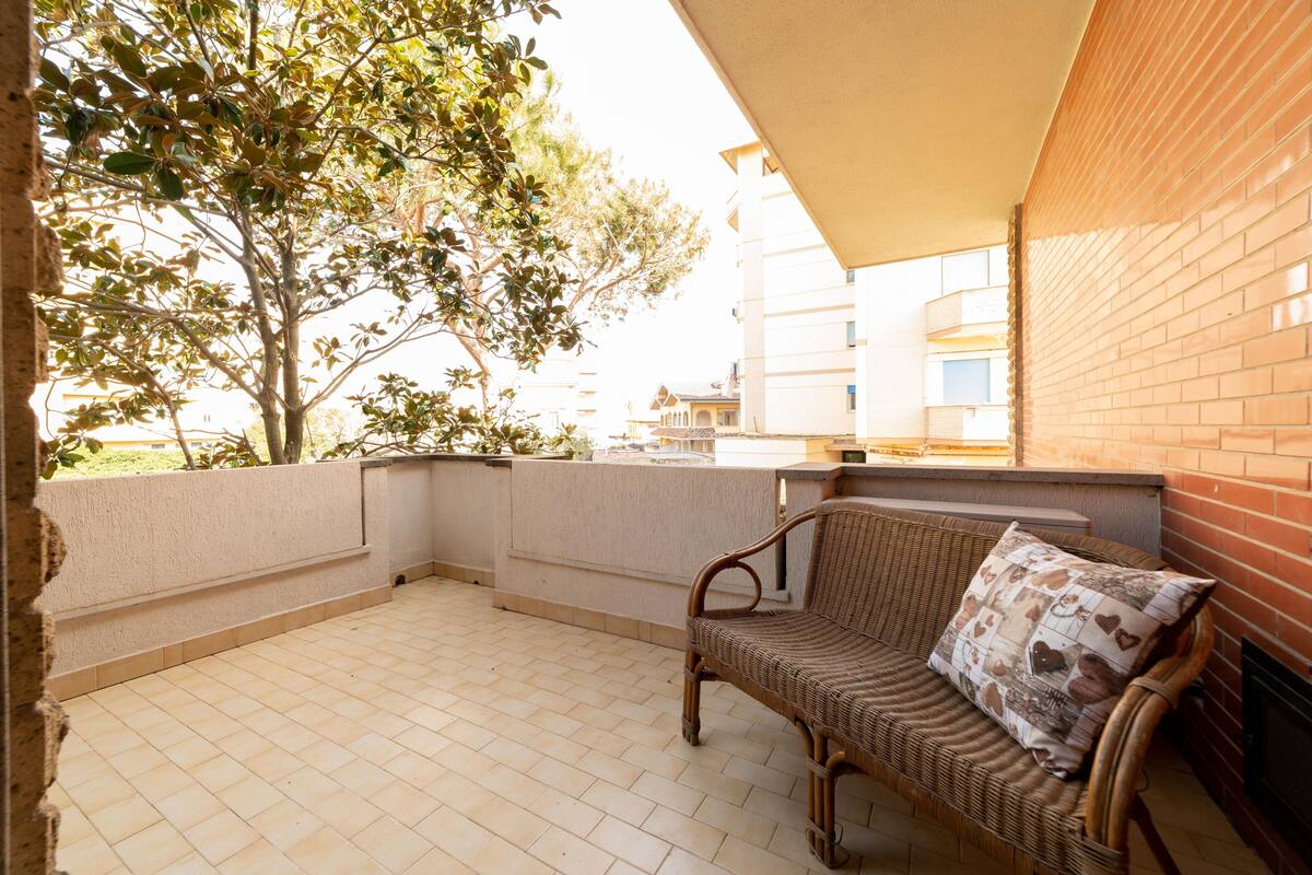 A balcony with a wicker sofa and a cushion, a tiled floor, and a view of buildings and trees.