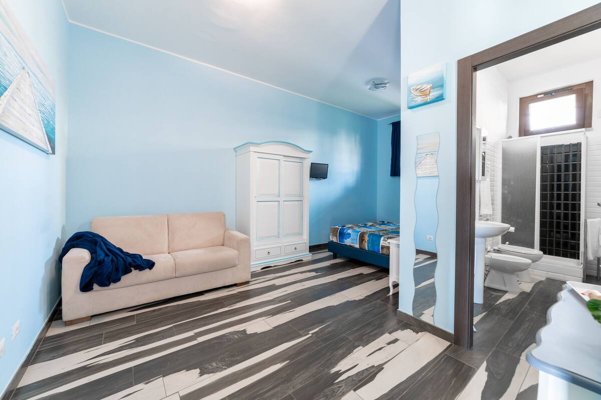A room with a beige sofa, a white cabinet, a bed with a blue headboard, a bathroom with a toilet and sink, and a patterned floor.