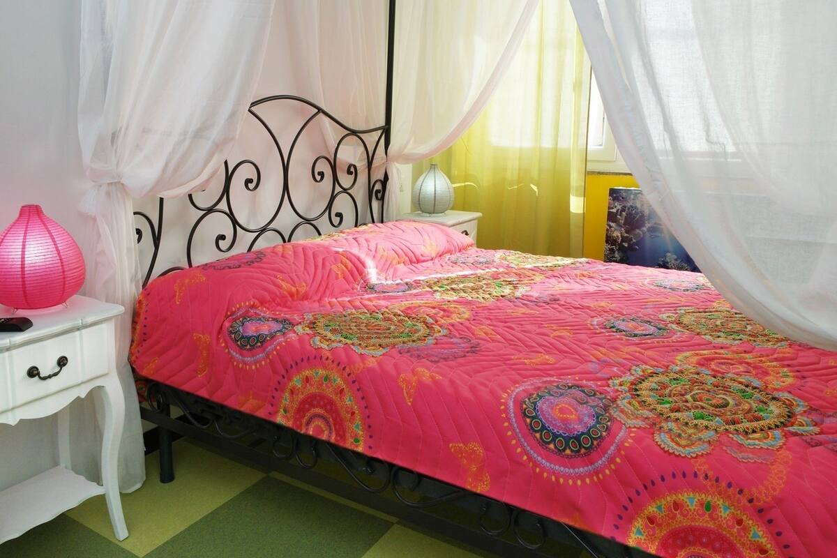 A bed with a pink quilt, a white bedside table, a pink lamp, and a black metal headboard.
