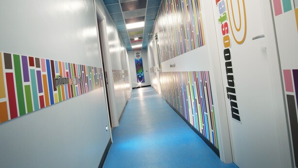 A brightly lit hallway with colorful vertical stripes on the walls, a blue carpet, and a ceiling with a grid pattern. - Milano Affittacamere (Milan)
