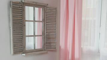 Una habitación con ventana, contraventanas de madera, cortina rosa y pared blanca.