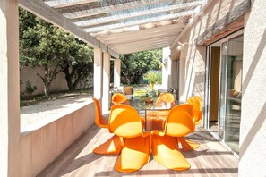 Une terrasse couverte avec une table en verre et des chaises orange, entourée d’un mur blanc et offrant une vue sur les arbres.