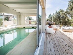 Un espace aménagé au bord de la piscine, avec une terrasse en bois, une piscine fermée par des parois de verre et des sièges d’extérieur.