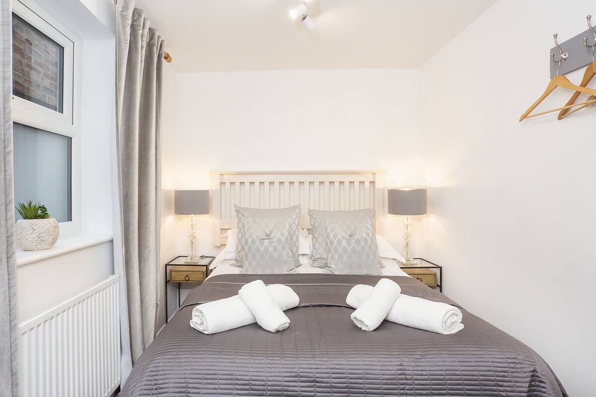 A neatly made bed with a grey quilt, two bedside tables with lamps, a window with curtains, and a wall-mounted coat rack.