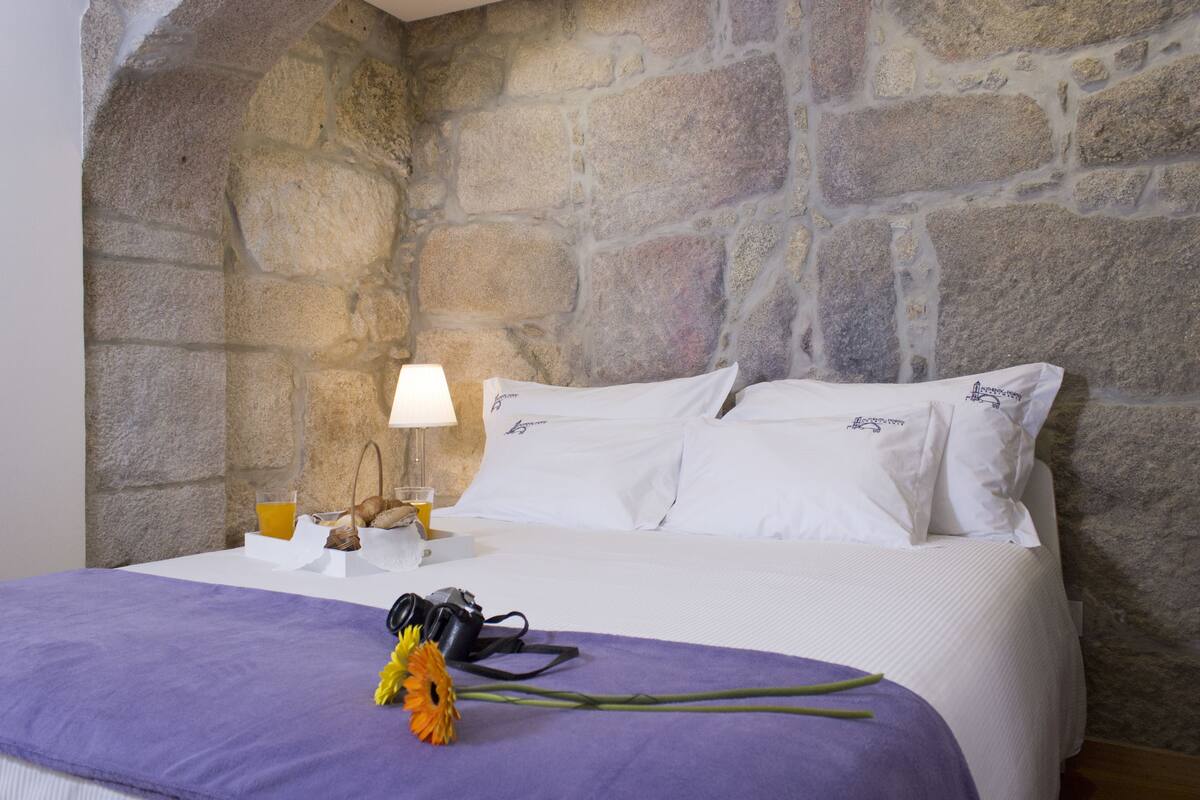 A bed with white linens, a purple throw blanket, a camera, and a bouquet of flowers on the bedside table.