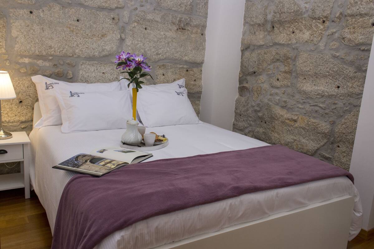 A neatly made bed with a purple blanket, white pillows, and a vase with flowers.