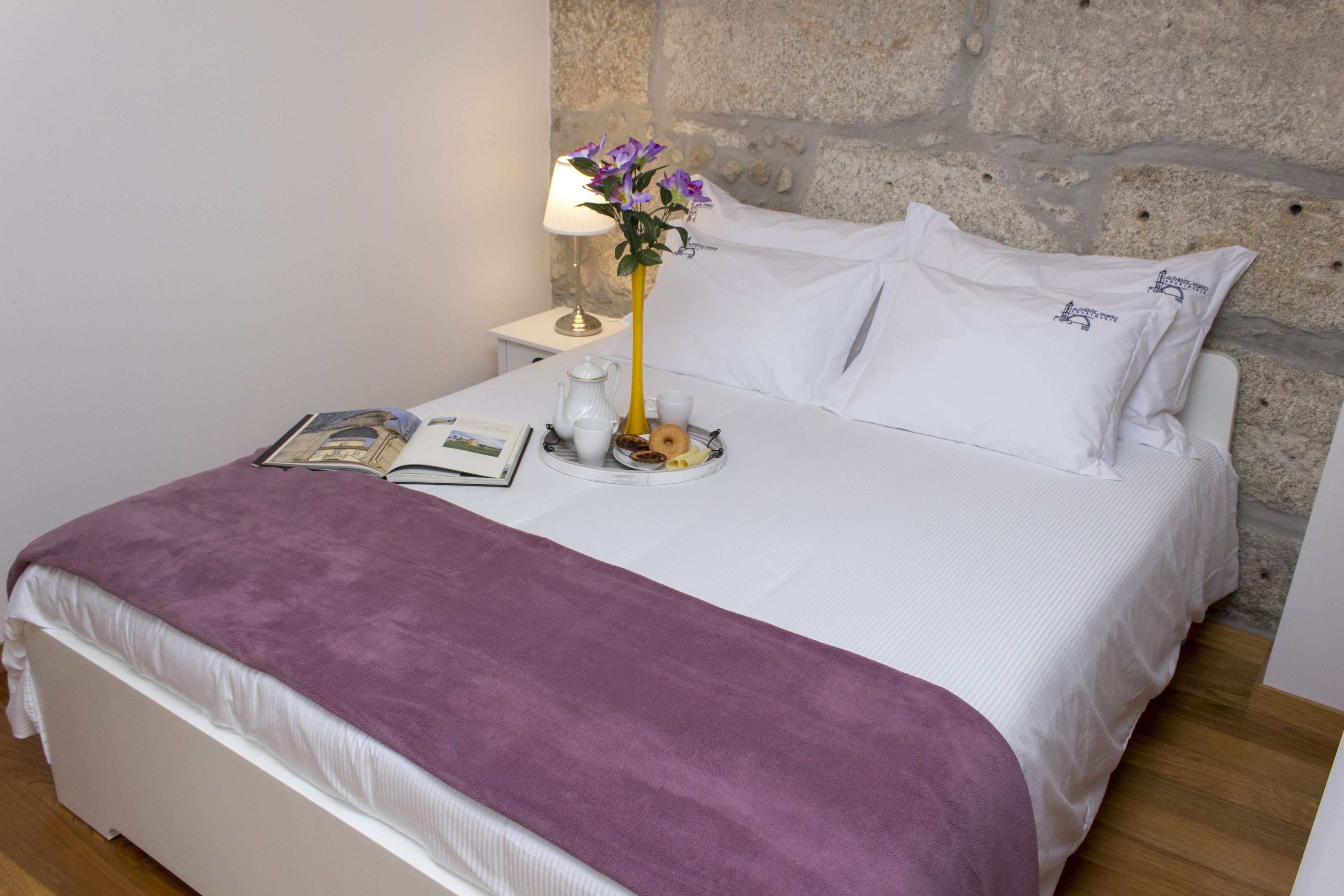 A neatly made bed with a purple blanket, a bedside table with a lamp and a book, and a stone wall behind.