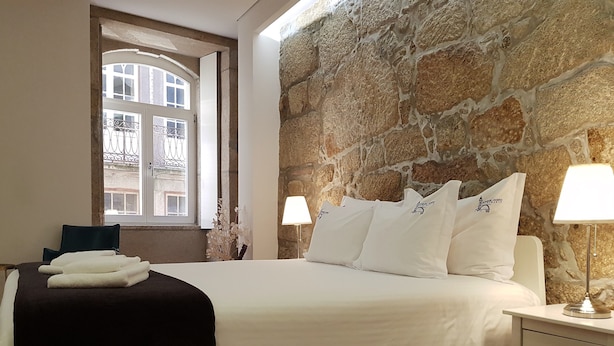 Una cama bien hecha con sábanas y almohadas blancas, una manta negra y una ventana con vistas a edificios. Hay dos lámparas de noche sobre una mesita.
