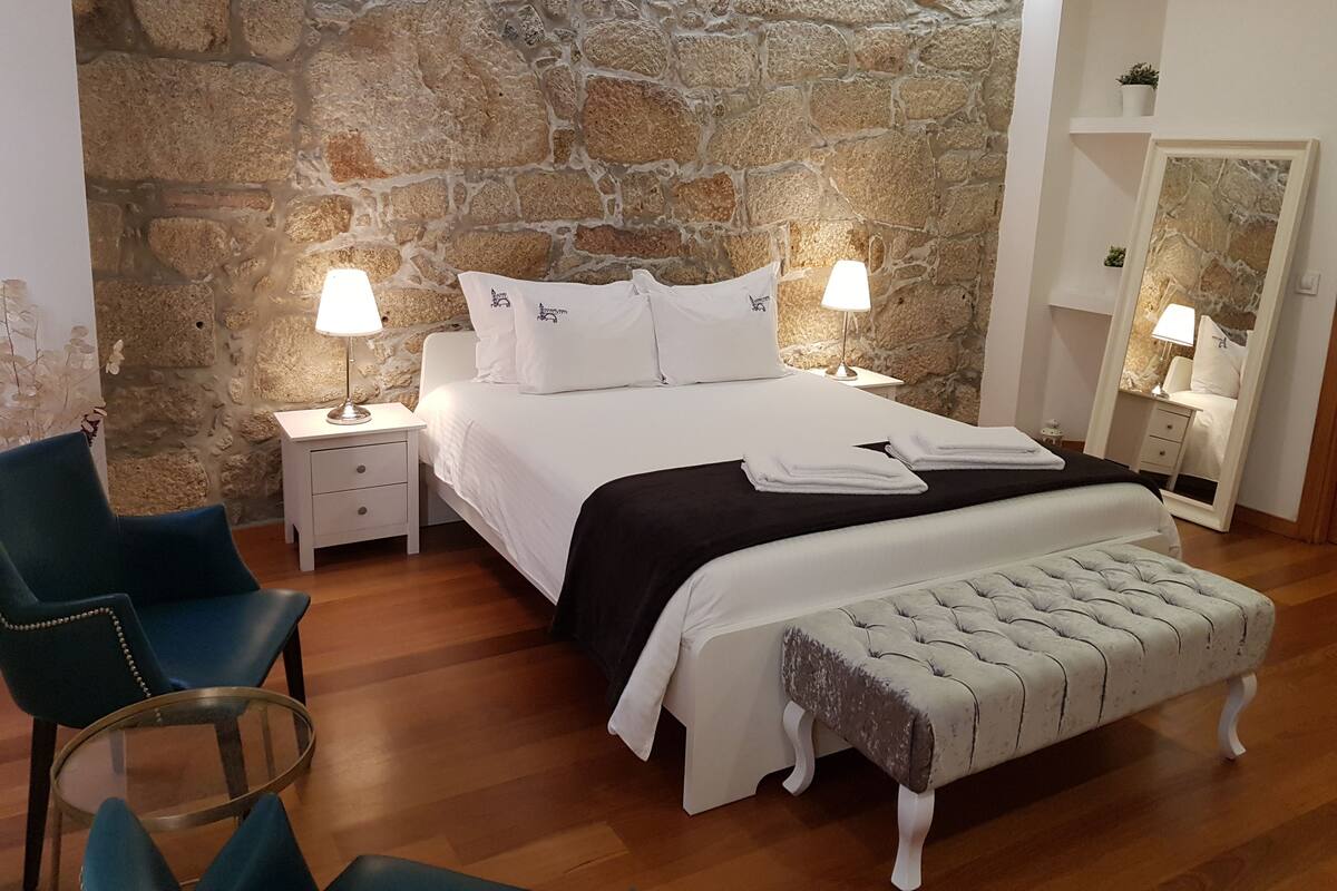A bedroom with a stone wall, a bed with white bedding, two bedside tables with lamps, a chair, and a bench.
