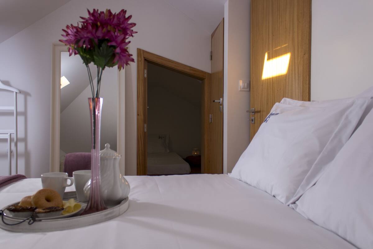 A neatly made bed with white linens, a tray with breakfast items, a vase with flowers, and a wooden door in the background.