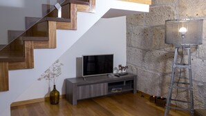 Una sala de estar moderna con una escalera de madera, un televisor sobre una consola gris, un jarrón con flores secas y una lámpara de pie.