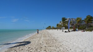 A wide sandy beach with clear blue water, palm trees, and a few buildings in the background. - White Villas (Providenciales)